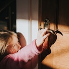 Cute little girl reaches up to open a door handle that is too high for her. She smiles knowingly at ...